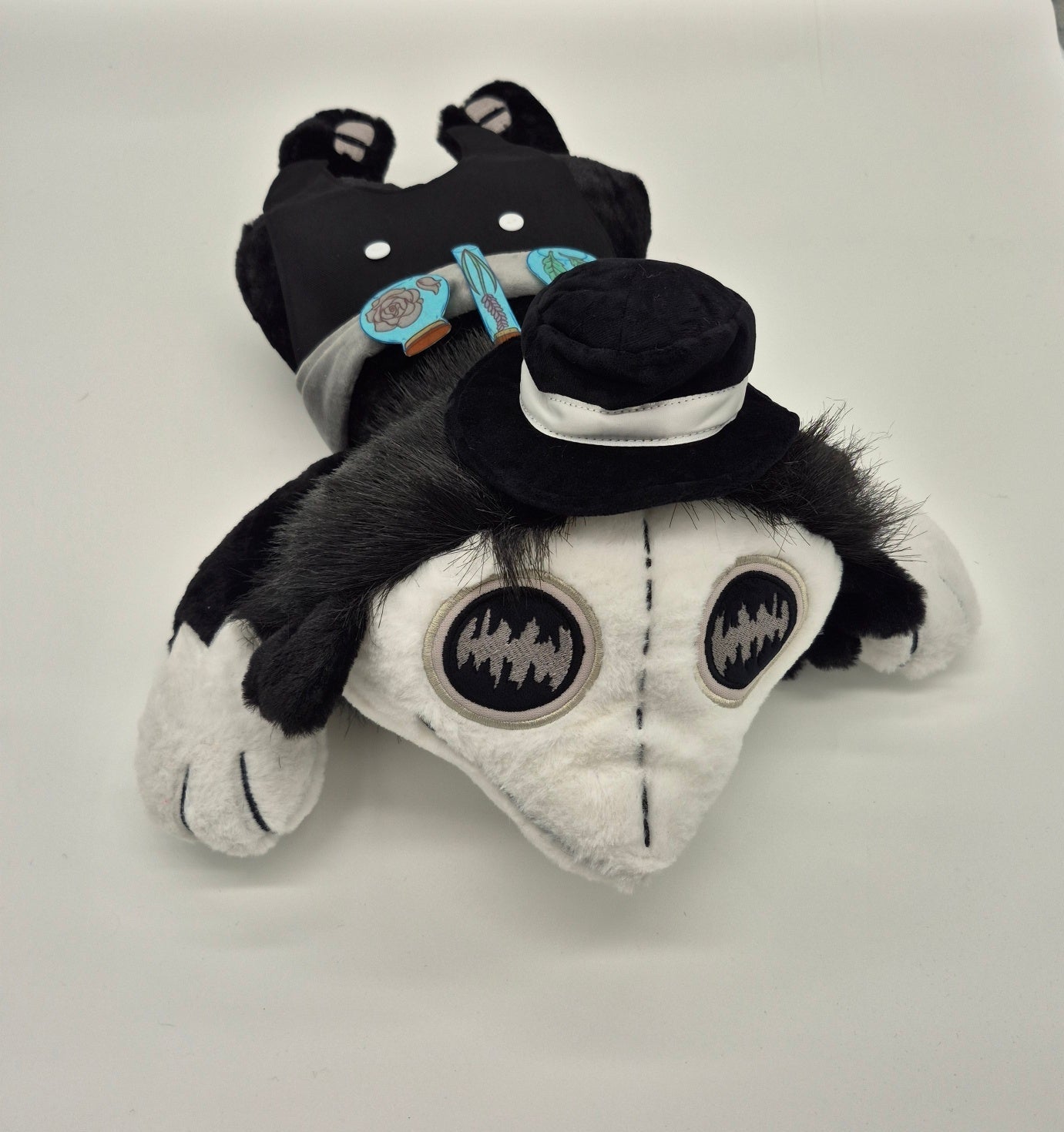 A plush toy shaped like a stylized plague doctor or creature with a black-and-white body, large circular stitched eyes, and a small black hat, photographed on a white surface with a metallic backdrop.