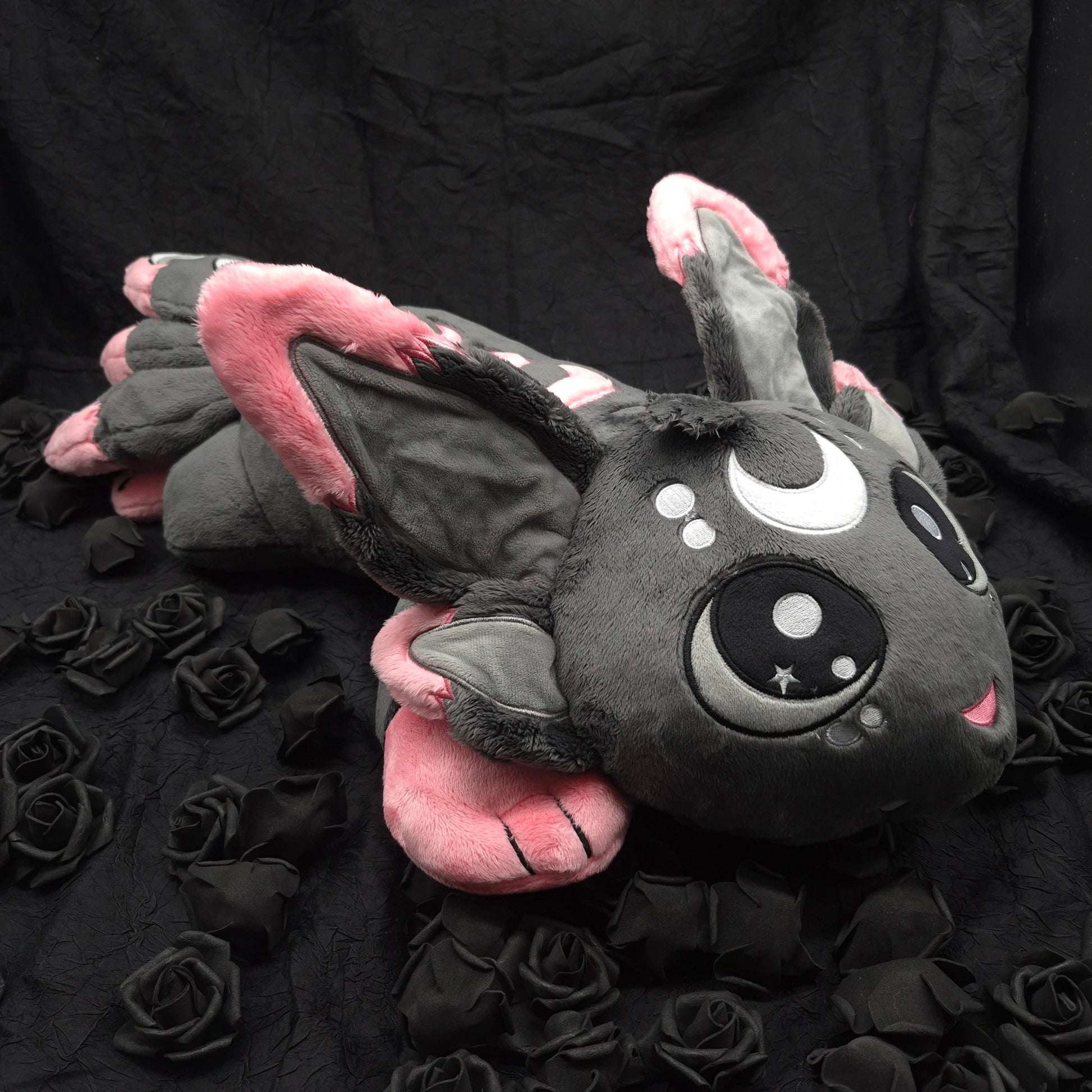 A dark gray plush bat toy with pink inner ears and wings, large cartoon-style eyes, and a small pink mouth, posed on a black fabric backdrop with black roses around it.