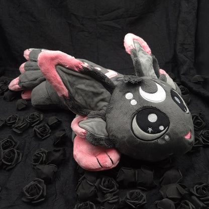 A dark gray plush bat toy with pink inner ears and wings, large cartoon-style eyes, and a small pink mouth, posed on a black fabric backdrop with black roses around it.
